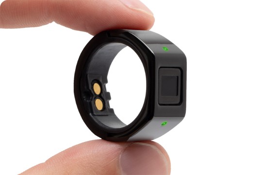 Hand holding a black smart ring with charging contacts and a small display.