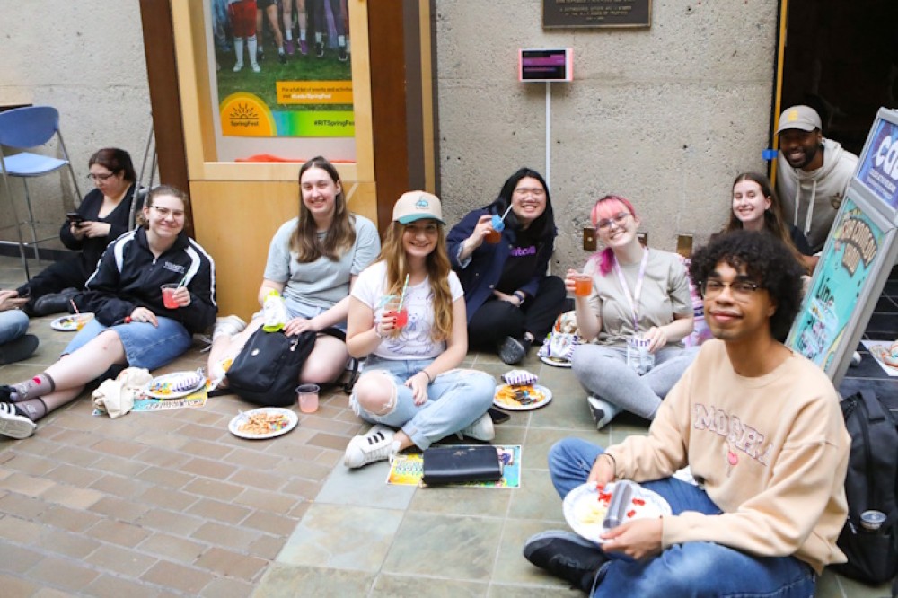 A group of students sit on the floor, enjoy a snack, and smile for the camera.