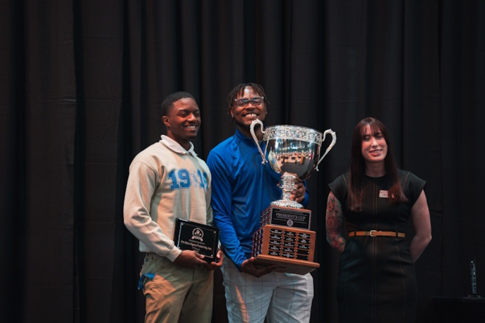 Two members of the Phi Beta Sigma fraternity hold up awards and smile for the camera–one holding a trophy and the other holding a plaque. An interpreter stands and smiles in the background.